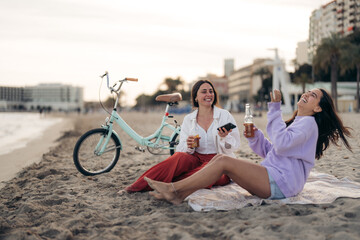 Happy young women with bike chilling at the beach, smiling, laughing, drinking beer, sitting on...