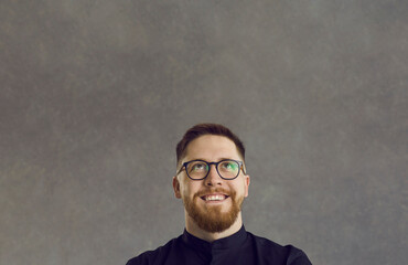 Smiling attractive young man with ginger beard looking up at blank space for text. Happy businessman or college student in geek glasses thinking of curious idea isolated on gray copy space background