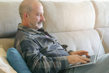 older man with moustache and plaid shirt sitting on the sofa at home with laptop computer