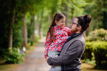 Fototapeta premium Young long hair bearded man in glasses standing with junior girl holding on hands on road in park