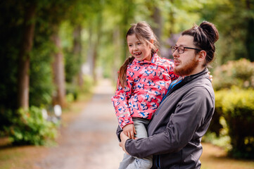 Fototapeta premium Young long hair bearded man in glasses standing with junior girl holding on hands on road in park