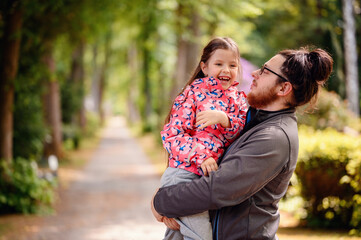 Fototapeta premium Young long hair bearded man in glasses standing with junior girl holding on hands on road in park