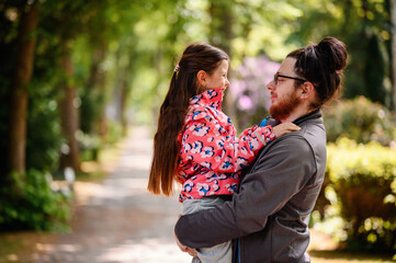 Young long hair bearded man in glasses standing with junior girl holding on hands on road in park