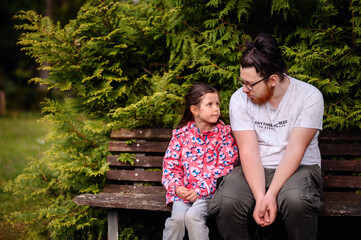 Young long haired man in glasses sits with junior girl on wooden bench