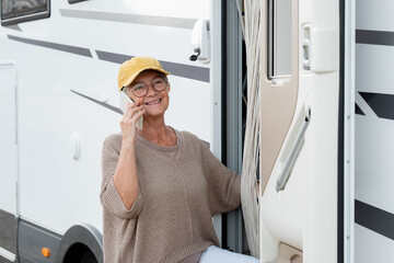 Happy senior woman standing outside a camper van motor home talking on mobile phone enjoying travel...