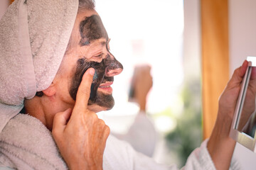 Cheerful beautiful senior woman in bathrobe applying a detox facial charcoal mask homemade looking...