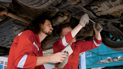 Two mechanic Caucasian man in large garage, Inspecting parts under raised car come in repair problematic part, male holding laptop in hand checking repairs on uncertain issues for correct solution.