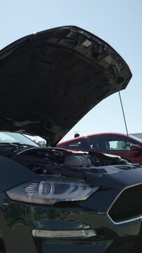 American muscle car stands with its hood up in open air parking lot. People are reflected in clean polished body, clean engine is demonstrated to passers by at meeting of automobile club.