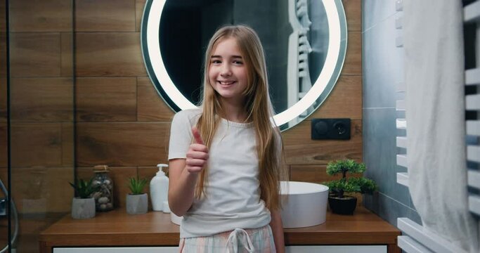 Portrait of smiling beautiful little girl in pajamas with long hair showing thumb up, looking at camera while standing in back of mirror at bathroom in home