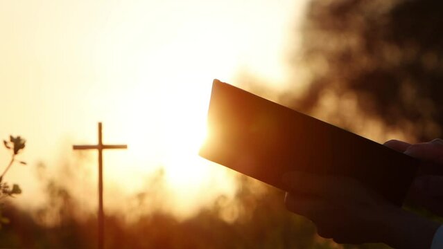 A Christian reading the Holy Bible in a beautiful evening with intense sunset light and a cross background symbolizing the death and resurrection of Jesus Christ
