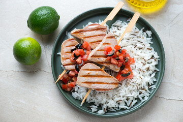 Green plate with bbq tuna skewers and mixed rice, horizontal shot on a light-beige stone background, high angle view