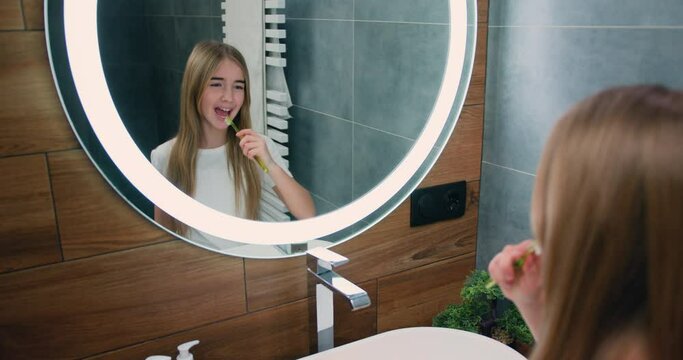 Cute caucasian little girl brushing teeth in front mirror after shower in the morning at bathroom. Healthy teeth, daily hygiene concept. Slow motion