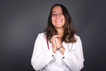 Dreamy charming young female with pleasant expression, keeps hands crossed near face, excited about something pleasant, poses against gray background.