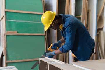 Male carpenter using hand saw cutting piece of wood during working in wood workshop. Male joiner wearing safety uniform, gloves, helmet and working in furniture workshop
