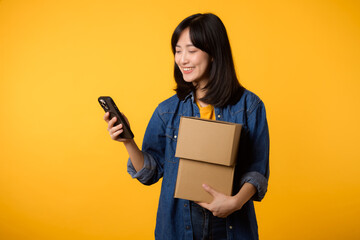 Portrait asian young woman wearing yellow t-shirt and denim shirt holding mobile phone and parcel box isolated on yellow studio background, Delivery courier and shipping service concept.