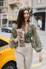 positive woman with long hair holding paper cup with coffee and newspaper while walking in trendy...