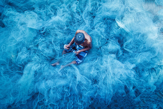 Old fisherman hands sewing blue fishing nets sitting on the ground and surrounded big net