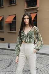 young woman with brunette long hair in beige pants, cropped blouse and handbag with chain strap standing with hand in pocket near blurred building on urban street in Istanbul