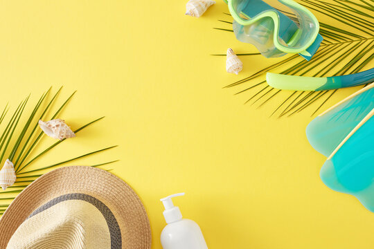 Seaside vacation concept. Top view flat lay of diving mask with snorkel, flippers, sunhat, cosmetic bottle, seashells and palm leaves on light yellow background with space for text