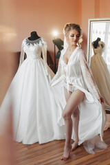 Naklejka premium A blonde bride in a long petticoat is standing in her room, posing near her wedding dress. Beautiful hairstyle, open legs. Voluminous dress, open bust
