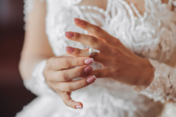 Blonde bride in long sleeve lace dress standing in her room, posing and putting on her engagement ring. Beautiful hair and makeup, open shoulders. Wedding portrait. French manicure
