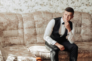 Portrait. A man wearing a white shirt, a black bow tie and black pants and a vest is posing in a room while sitting on a sofa. A stylish watch. Men's style. Fashion. Business