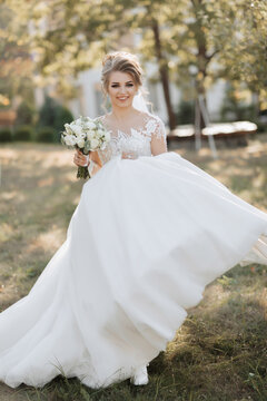 Wedding portrait. A blonde bride in a white dress with a train is walking, smiling and holding a bouquet and her wedding dress. Photo session in nature. Sun rays in the photo