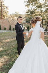 Wedding portrait. A groom in a black suit and a blonde bride walk holding hands in a garden. Rear view. A white, long veil in the air. Photo session in nature. Beautiful hair and makeup