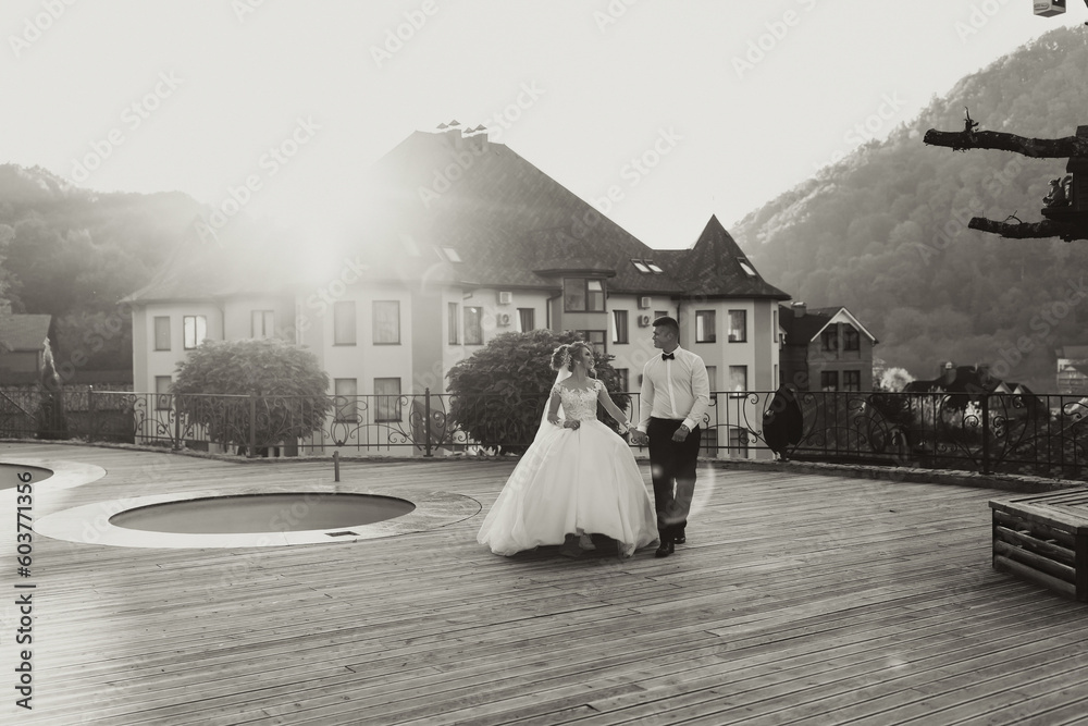 Wall mural Wedding black and white portrait. The groom in a black suit and the blonde bride are walking on the background of the mountain and buildings, near the pool. Long dress in the air. Romantic photo