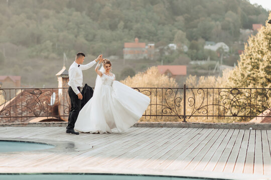 Wedding portrait. A groom in a black suit and a blonde bride dance a waltz against the background of a mountain and buildings, near a swimming pool. Long dress in the air. Romantic photo