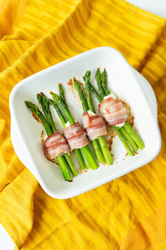 Asparagus baked with bacon and various spices. A bright background, an orange napkin on which the dish stands. Vertical photo.