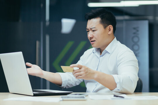 Problems with the transaction. Angry young Asian businessman sitting in office in front of laptop, looking worriedly at monitor, holding credit card in hand.