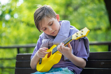 a boy plays a small guitar, a yellow ukulele on a bench in the park