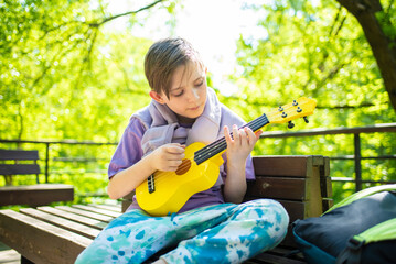 a boy plays a small guitar, a yellow ukulele on a bench in the park	