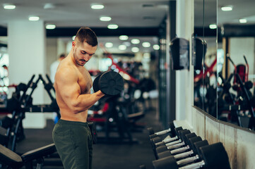 Fototapeta na wymiar A strong muscular bodybuilder is working out with a dumbbell and looking at his biceps flexing at the gym.