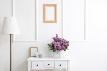 Vase with lilac flowers, candles and frame on table in light room