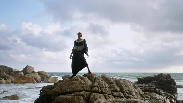Androgynous person of color stands in boutique posh dress on rock above ocean coastline. Trans sexual ethnic fashion model in epic outfit wears golden jewellery, posing like Oshun goddess concept.