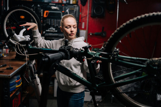Wide shot of pretty blonde cycling mechanic female watching rear shifter of mountain bike, changing speeds using handlebar shift lever, working in bike repair workshop with dark interior.