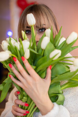 Portrait of a young girl with blond hair smelling white tulips holding bouquet with hands in room inside 