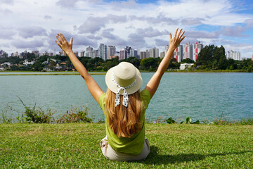 Young woman with hat raising arms sitting on grass in Barigui Park, Curitiba, Brazil