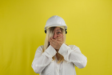 On an isolated yellow background, a woman in a construction helmet funny closes her mouth. A woman engineer in a funny fright holds her mouth with her hands. Engineer funny emotion concept.