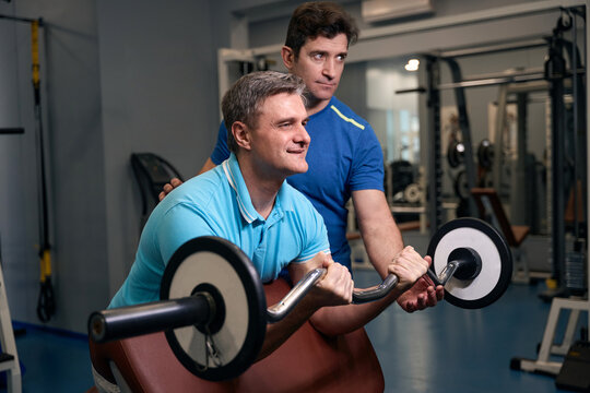 Pleased adult male performing bicep strength exercise with personal trainer