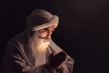 A senior man in a turban is associated with a Muslim standing in dark smoky background light ray...