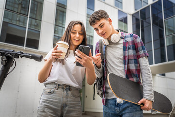Two teenagers with headphones and skateboard looking at mobile phone and having fun, modern...