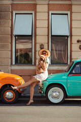 Young woman sitting on the hood of a retro car on a summer day playing with her puppy