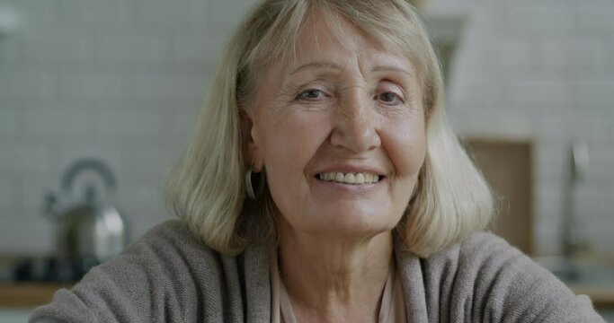 Close-up portrait of beautiful elderly lady with blond hair smiling looking at camera in kitchen in apartment. Retirement and happy senior people concept.