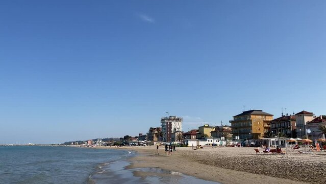 Intriguing panorama in Porto San Giorgio on the Adriatic Sea with a paraglider in the sky above the beach circling a black and white building with different onlookers, parasols and sun loungers