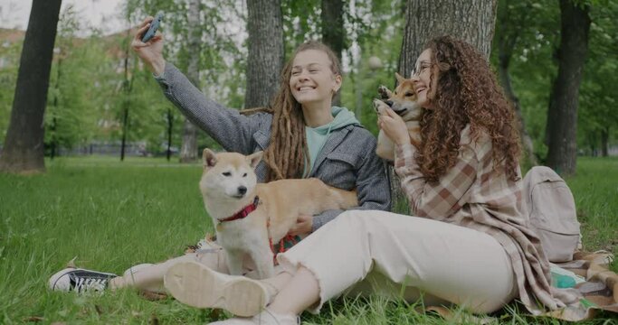 Young women friends taking selfie with shiba inu dogs posing for smartphone camera having fun with pets in park. Friendship and animals concept.