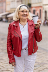 Senior woman enjoying morning coffee hot drink and smiling outdoors. Relaxing, taking a break. Elderly grandmother walking in urban city center street, drinking coffee to go. Town lifestyles. Vertical