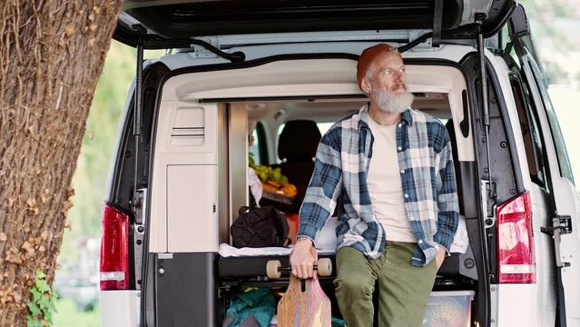 Active cool old hipster man standing near rv camper van holding skateboard. Mature traveler skater traveling in campervan enjoying camping, freedom spirit and extreme activities in nature park.